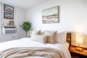 A neatly made bed with a white and beige blanket, a bedside lamp, and a wall-mounted air conditioner. - Rare Corner Stunning Ocean View 30F -2 Units A + B (Honolulu)