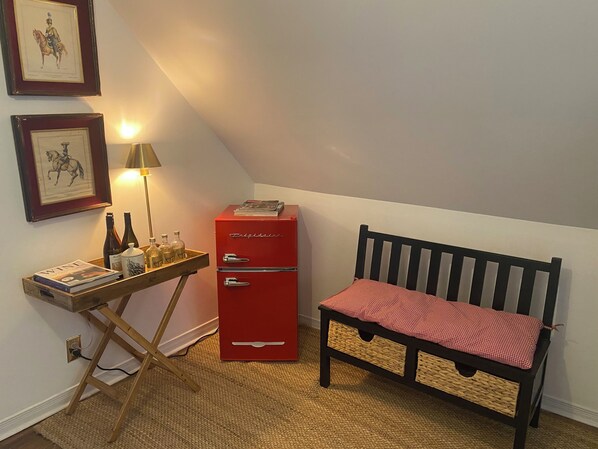 Ein gemütliches Zimmer mit einem roten, alten Kühlschrank, einem Holztischchen mit Weinflaschen und einer Lampe, einer rot-weiß karierten Bank und gerahmten Kunstwerken an der Wand.