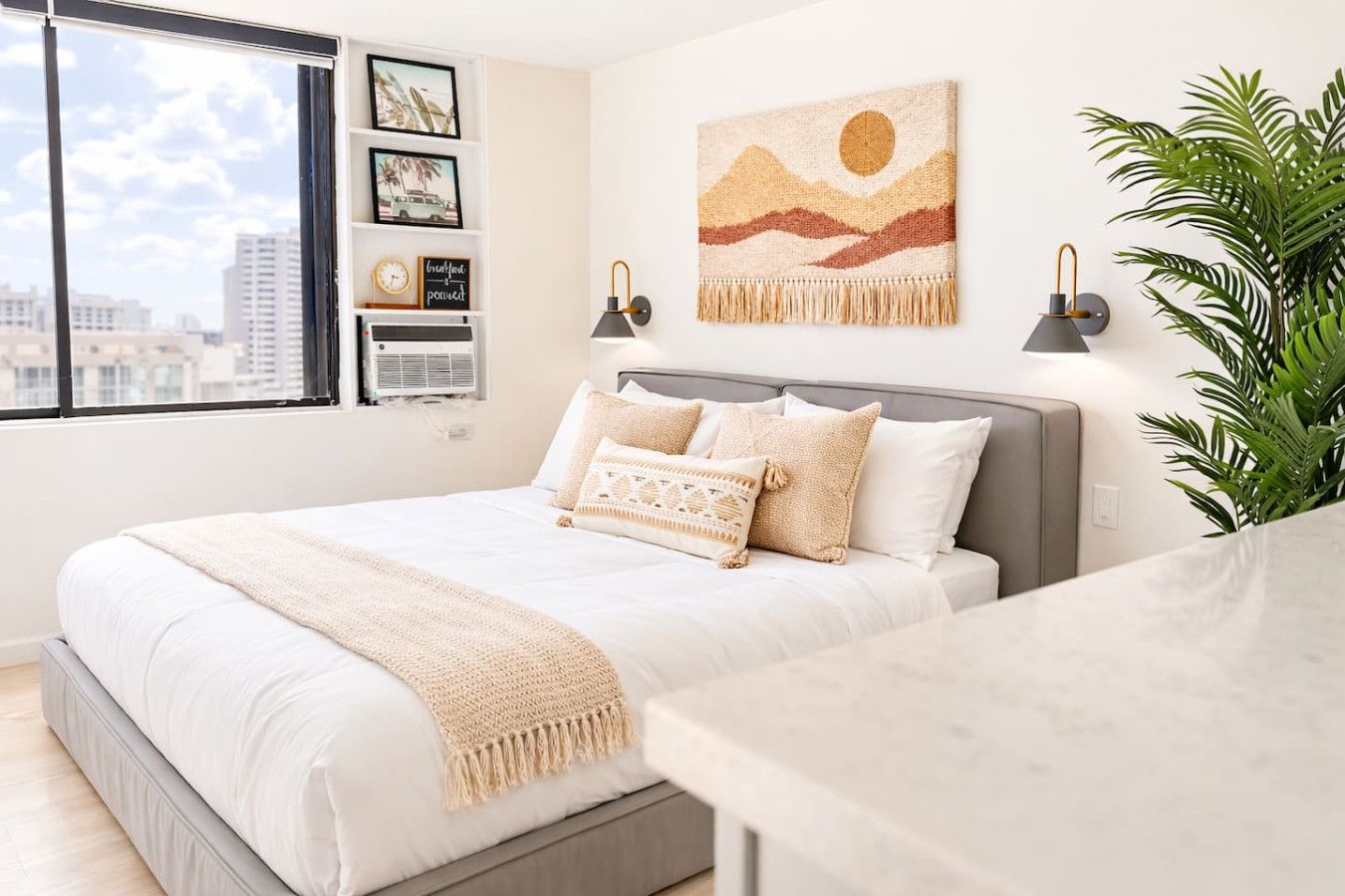 Un dormitorio con cama, mesita de noche, lámpara de pared, planta y vista panorámica de la ciudad a través de una ventana.
