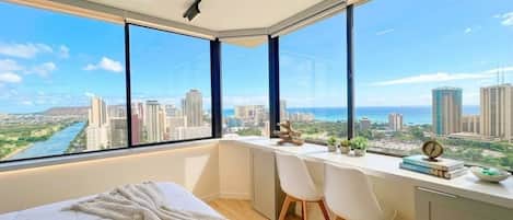 Un dormitorio con un ventanal grande, una cama, un escritorio y vistas a un paisaje urbano.