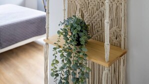 Une décoration murale en macramé avec des étagères, une plante en pot et une plante suspendue, dans une pièce comprenant un lit et une fenêtre.