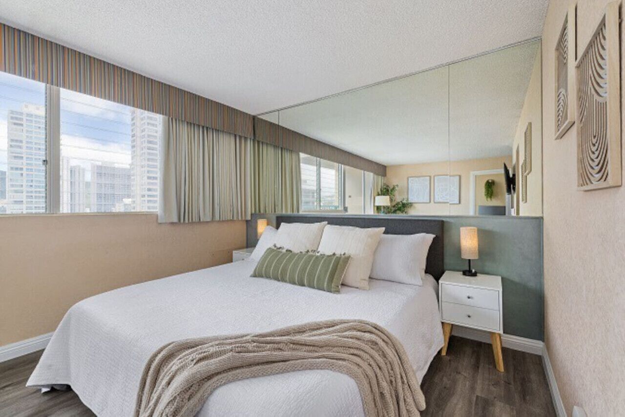 A modern hotel room with a large bed, a nightstand, a mirror, and a view of the city.