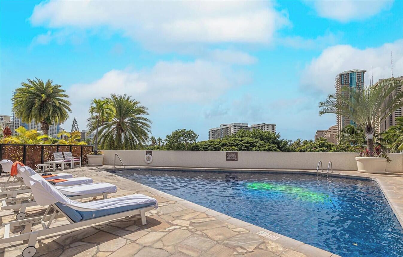A swimming pool with lounge chairs, palm trees, and high-rise buildings in the background.