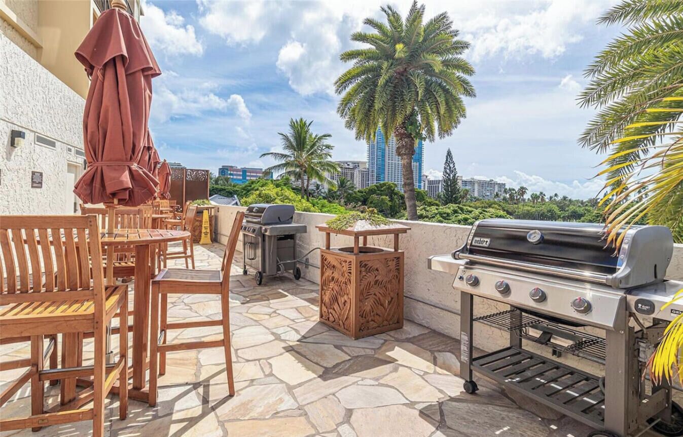 An outdoor patio with a grill, seating, and a view of palm trees and buildings.