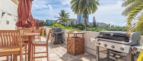 An outdoor patio with a grill, seating, and a view of palm trees and buildings.