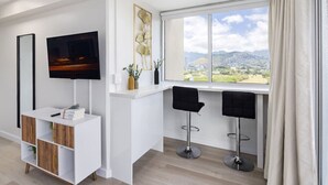 Ein modernes Wohnzimmer mit Fernseher, weißem Lowboard, zwei Barhocker und einem großen Fenster mit Blick auf die Stadt.