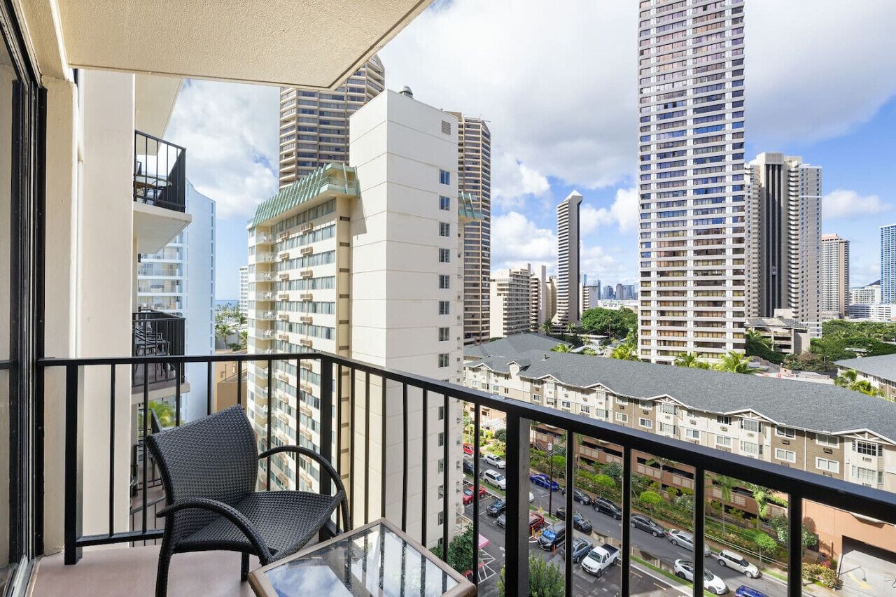 A balcony with a chair and a table, overlooking a cityscape with tall buildings and a parking area.
