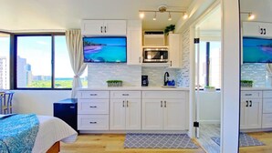 A modern kitchen with white cabinets, a microwave, and a coffee maker. There is a large window with a view of buildings and a bed with a blue blanket in the room. - HM15 Rare Find - Corner Ocean View Studio at Waikiki (Honolulu)