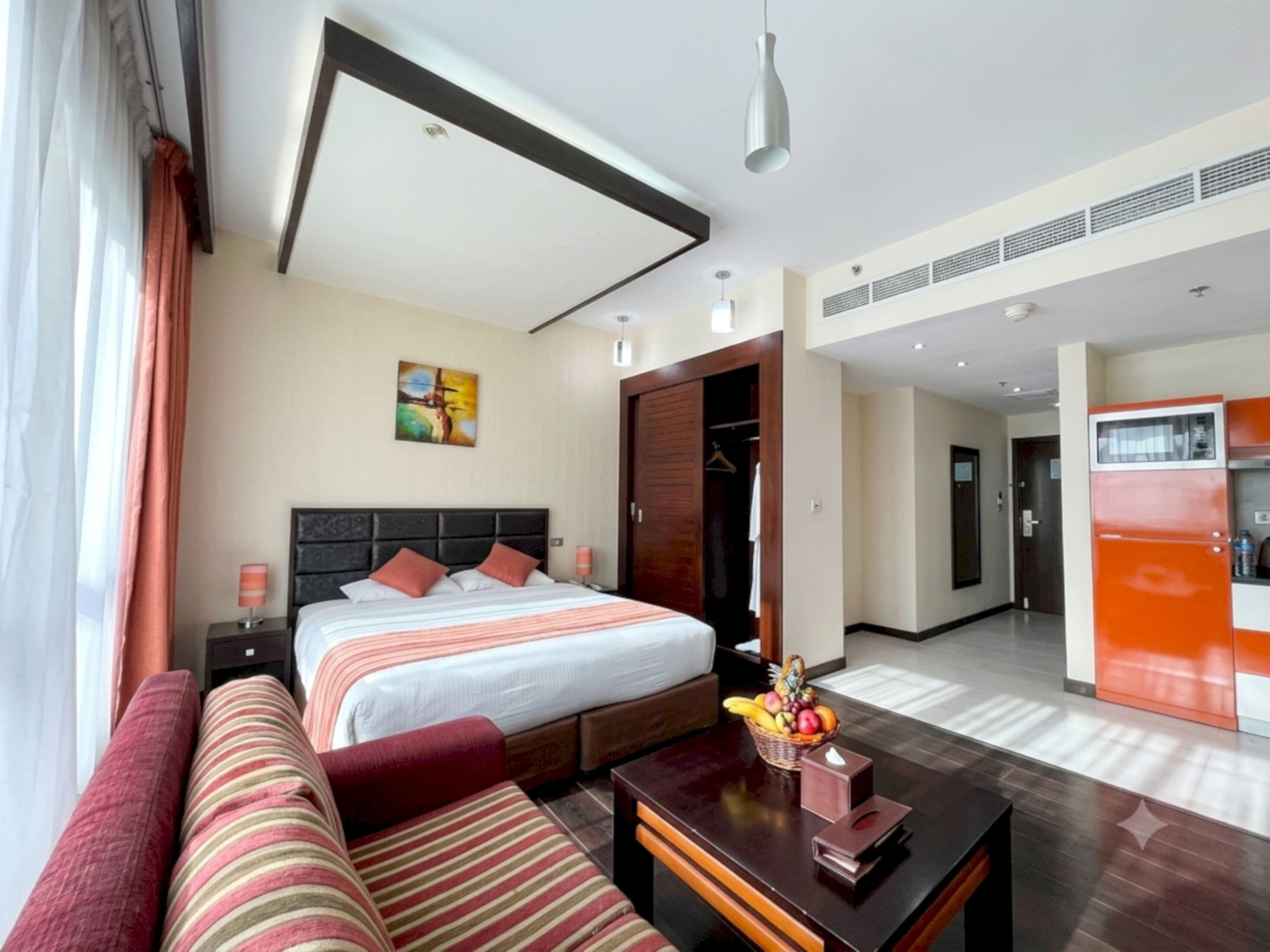 A hotel room with a bed, a sofa, a desk with a fruit basket, a wardrobe, and a kitchenette.