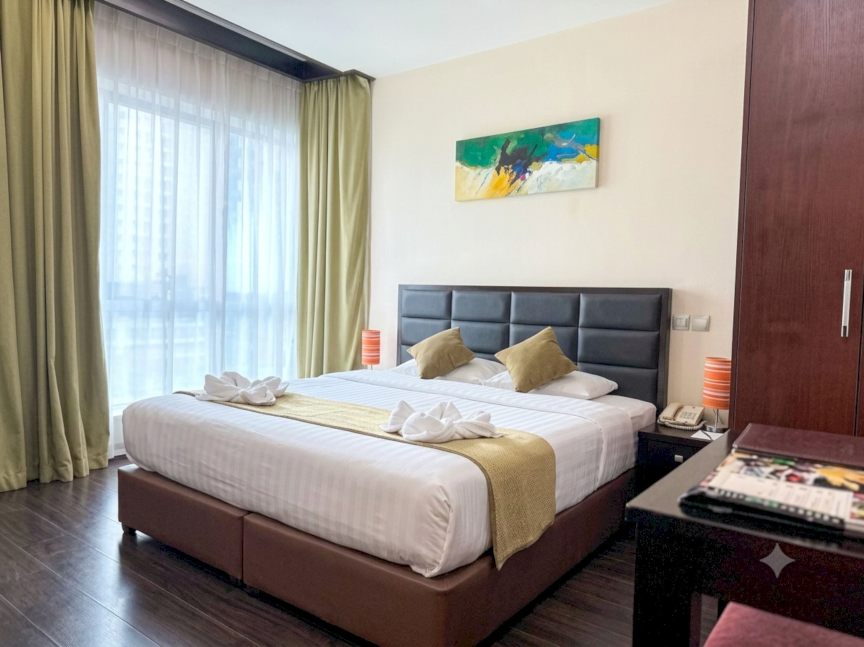 Una habitación de hotel con una cama grande, cabecero, un cuadro en la pared, escritorio con lámpara y armario.