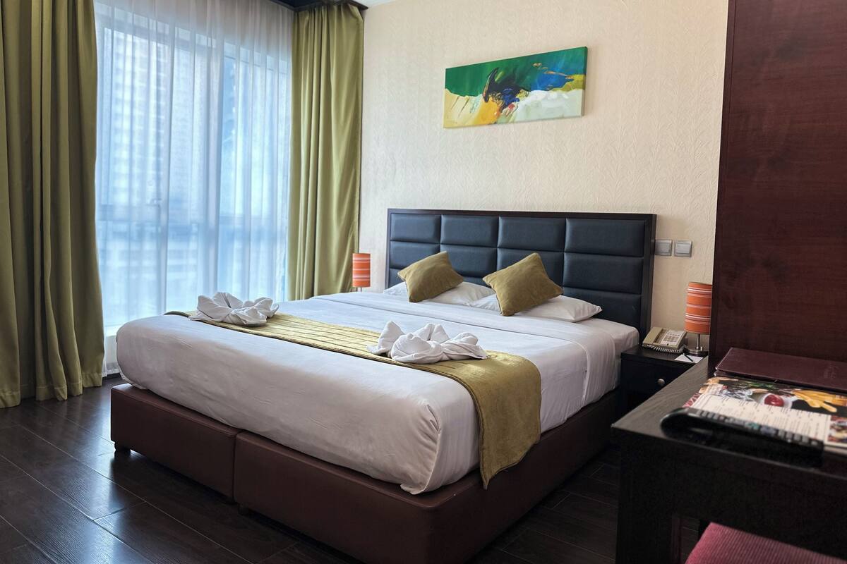 Ein Hotelzimmer mit einem großen Bett, einem Kopfteil, einem Bild an der Wand und einem Fenster mit Vorhängen.