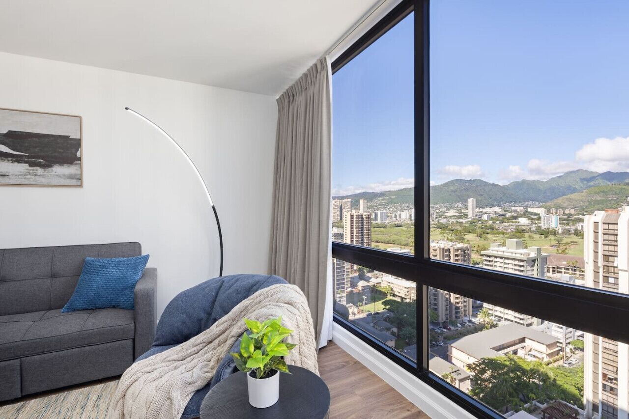 Una sala de estar moderna con un sofá gris, una almohada azul, una manta blanca y una planta en maceta sobre una mesita. Una lámpara de pie está colocada cerca del sofá. La habitación tiene un ventanal con vistas a un paisaje urbano y montañas.