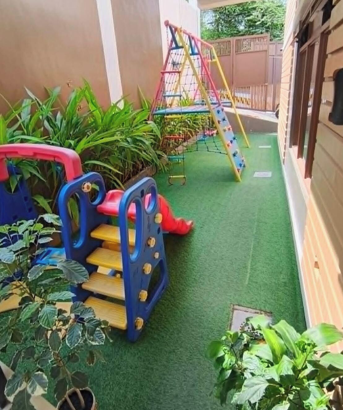 A children's play area with a slide and climbing net, surrounded by green plants.