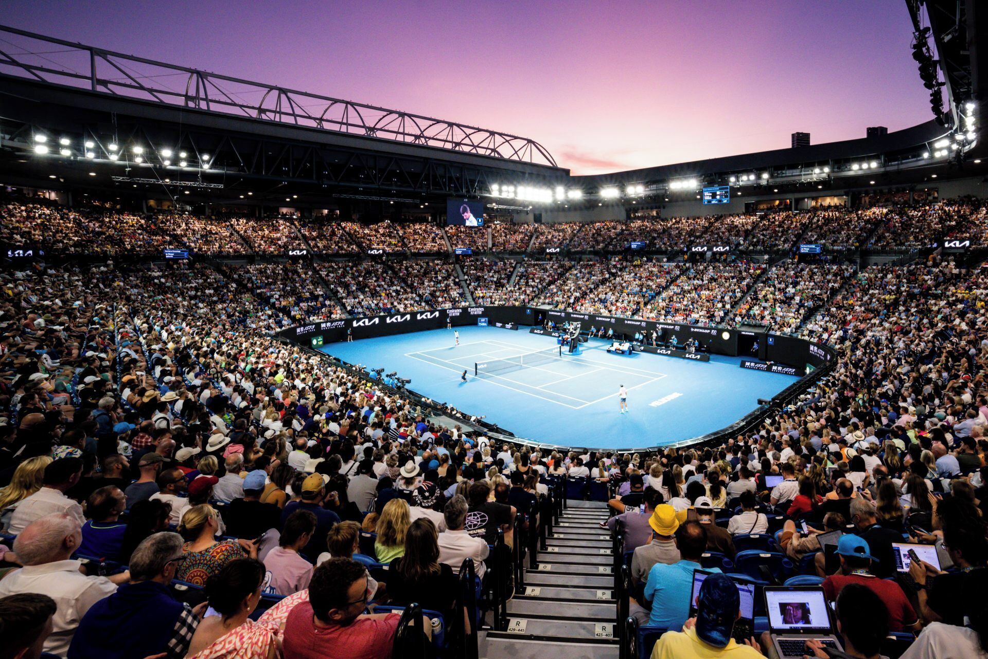 En stor indendørs arena med flere niveauer af siddepladser, en central tennisbane og et højtloftet rum med synlige konstruktionsbjælker.