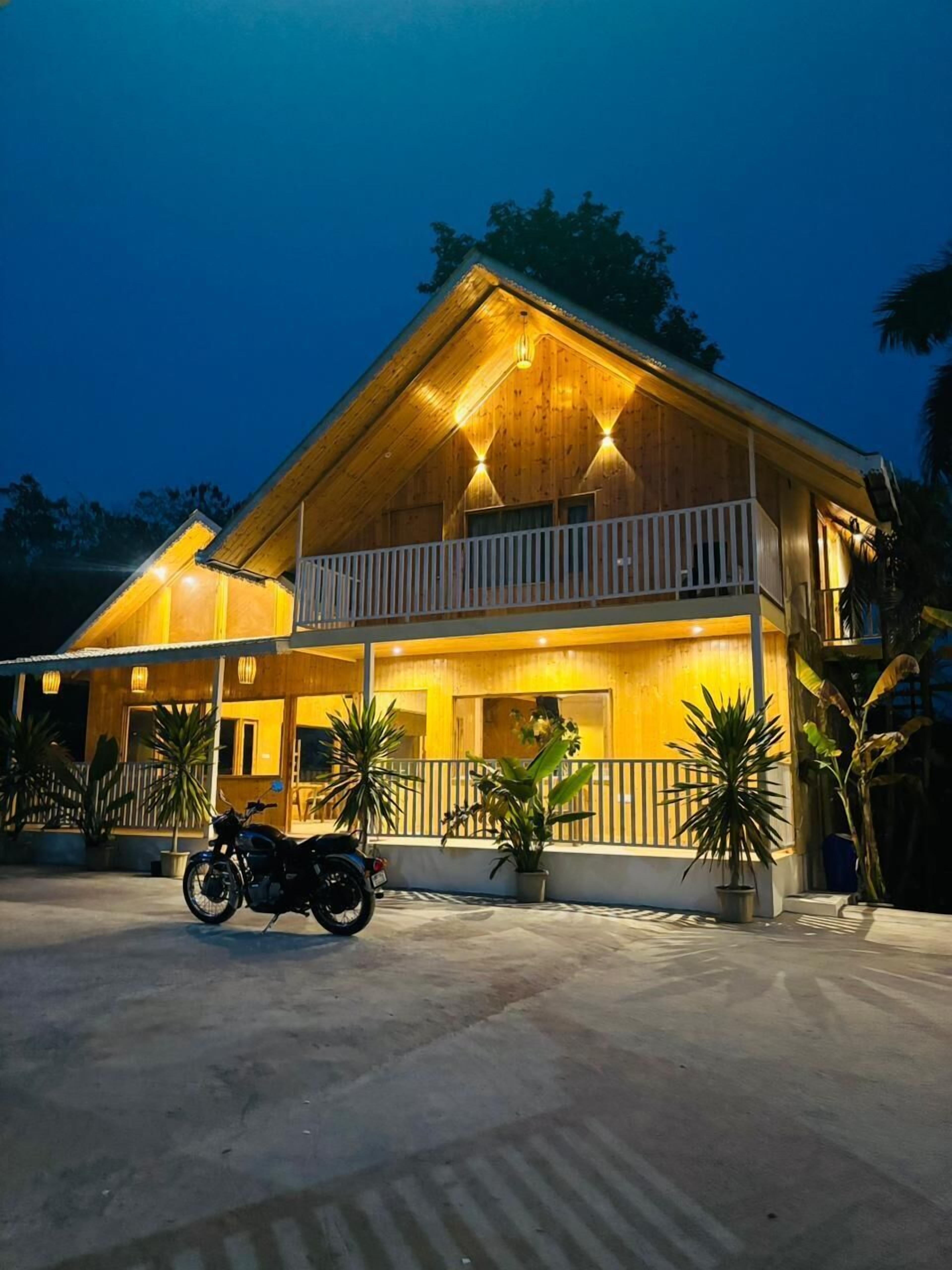 A two-story wooden house with a balcony, potted plants, and a motorcycle parked in front.