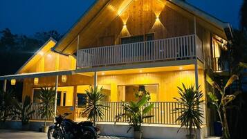 A two-story wooden house with a balcony, potted plants, and a motorcycle parked in front.