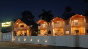 A row of wooden cabins with balconies, a sign for 'BIRDELA WOODS' indicating a guesthouse and restaurant, and a white fence with lights. - Bundela Woods Cottage and Restaurant (Rajnagar)