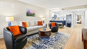 Een moderne woonkamer met een bank, twee fauteuils, een salontafel en een vloerkleed. Er is een keukenhoek met een bar en een koelkast.