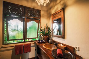 Un baño con tocador de madera, un ventanal con vistas a la vegetación, una planta en maceta y un espejo.