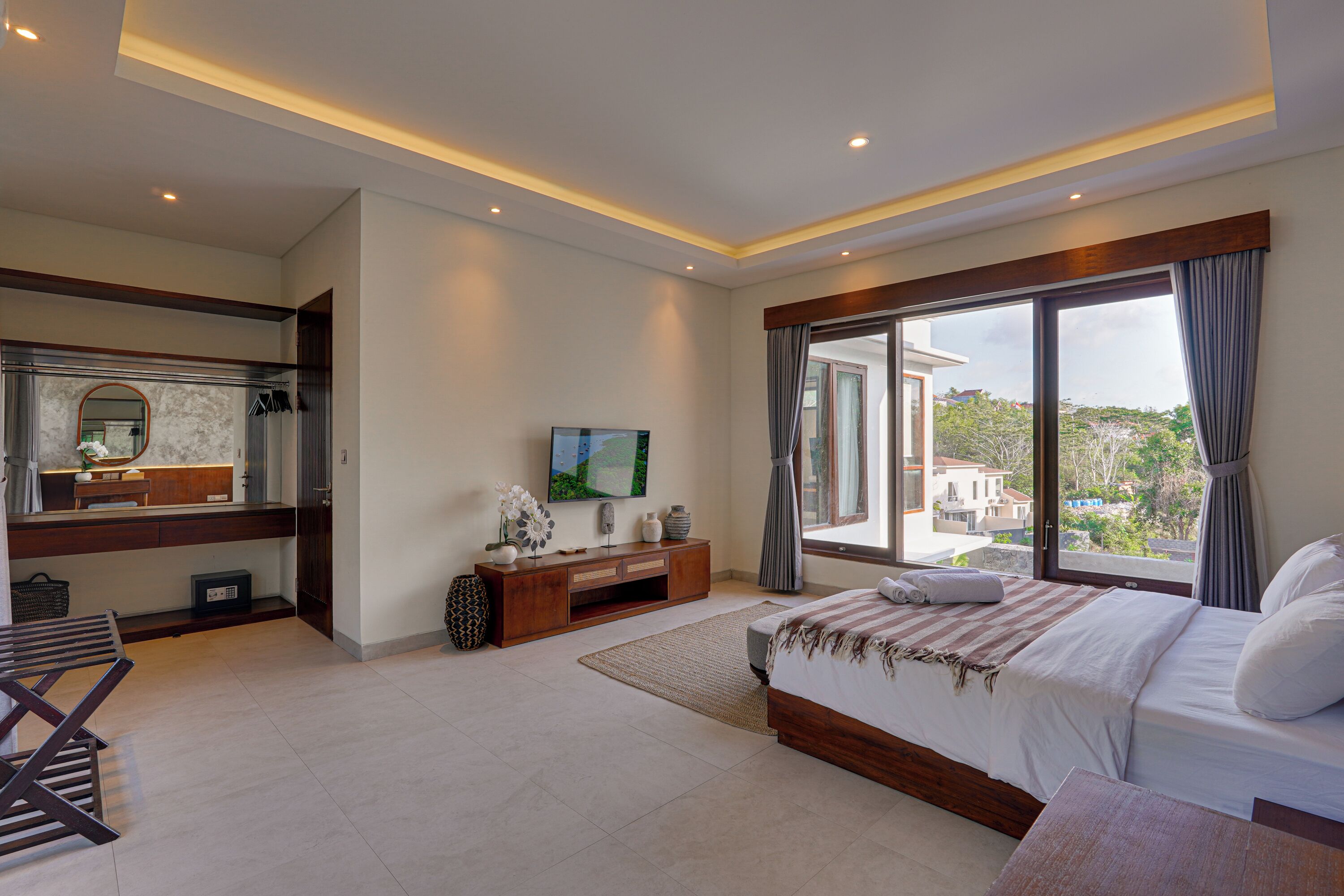 A modern bedroom with a large bed, a TV, a vanity, and a balcony with a city view.