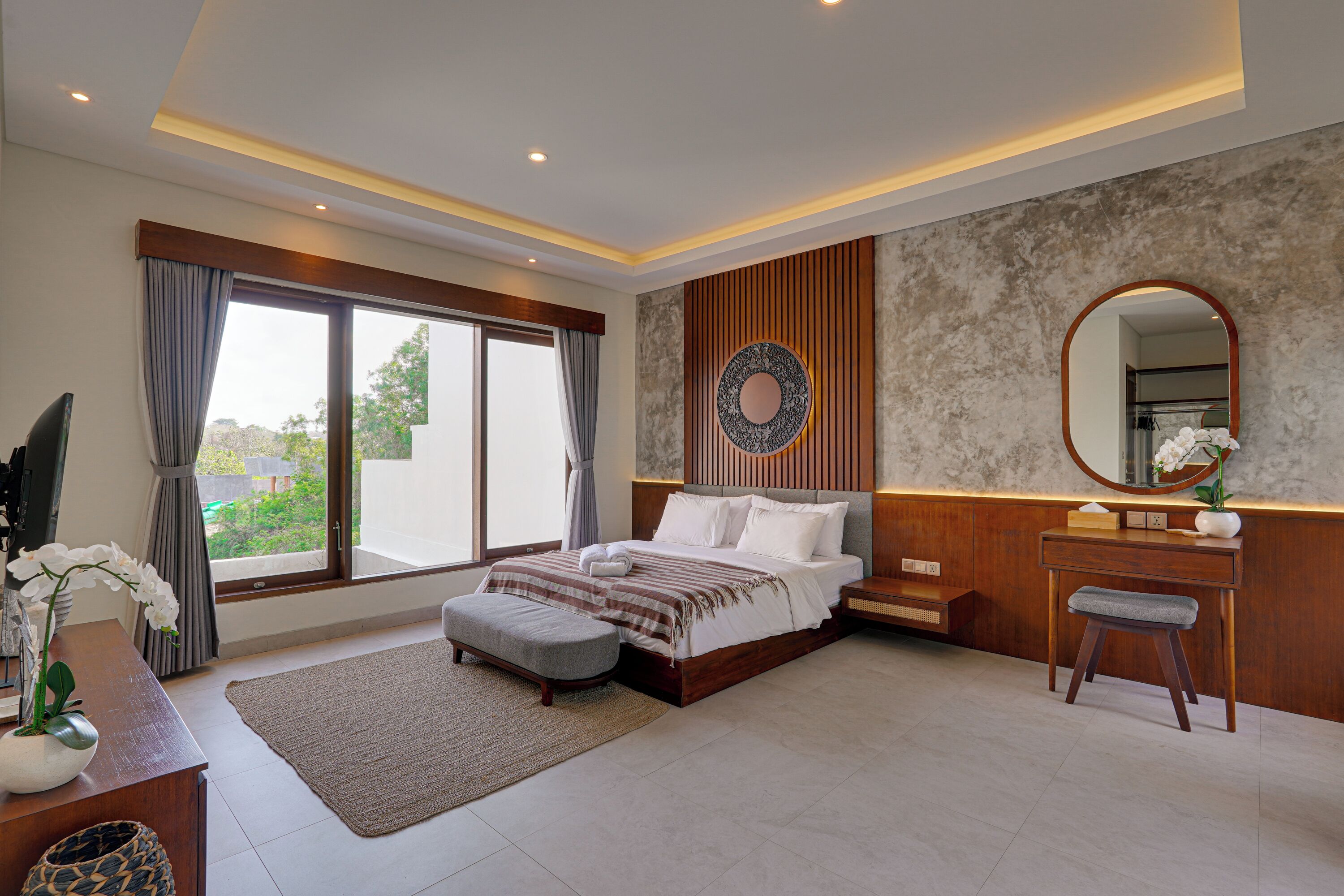 A modern bedroom with a large bed, a vanity with a mirror, and a view of greenery outside.
