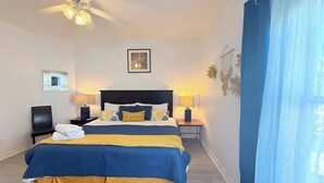 Quarto com cama, duas mesas de cabeceira, ventilador de teto, um quadro na parede e janela com cortinas.