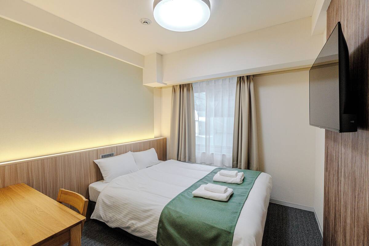A hotel room with a bed, a desk, a chair, a flat-screen TV, and a window with curtains.