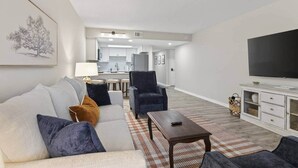 A modern living room with a sofa, armchairs, a coffee table, and a TV. There is a kitchen in the background with white cabinets and a dining area with stools. - Charming 2BR/2BA Condo - Steps from Auburn Campus (Auburn)