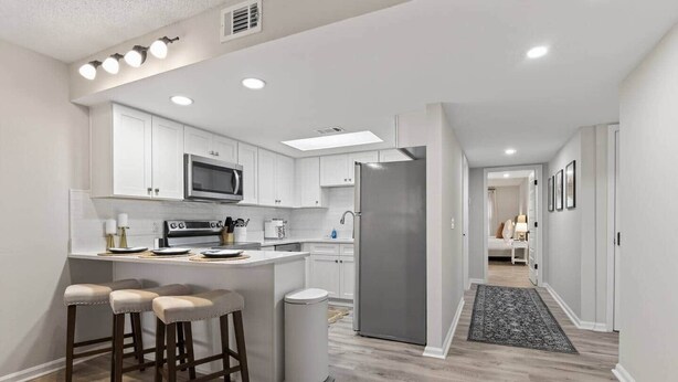 A modern kitchen with white cabinetry, a central island with stools, a microwave, and a refrigerator. - Charming 2BR/2BA Condo - Steps from Auburn Campus (Auburn)