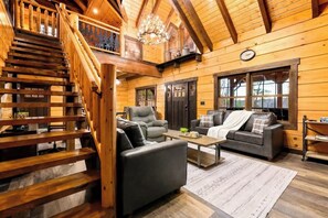 A log cabin interior with a wooden staircase, a living room with a sofa, armchair, and coffee table, and a dining area with a table and chairs. - Luxury Chalet Retreat with Prime Location, Hot Tub, Pond (Sevierville)