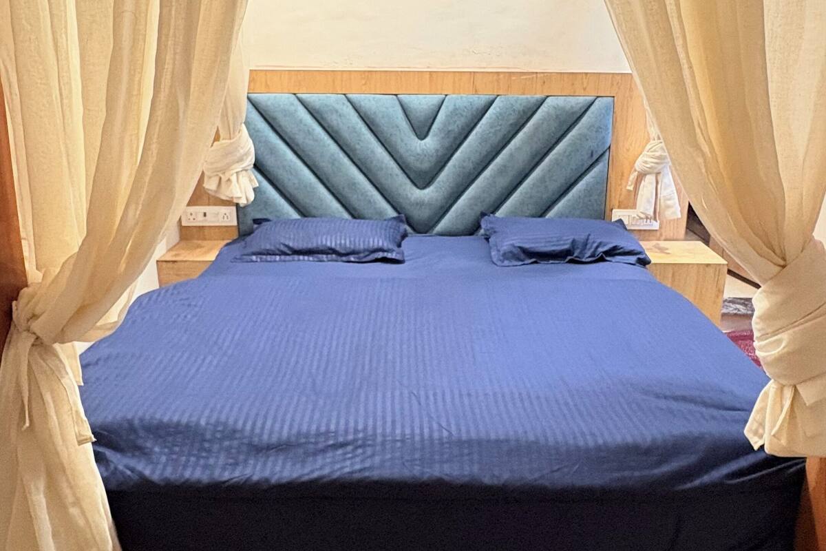 A bed with a blue bedspread, a wooden headboard, and sheer curtains.