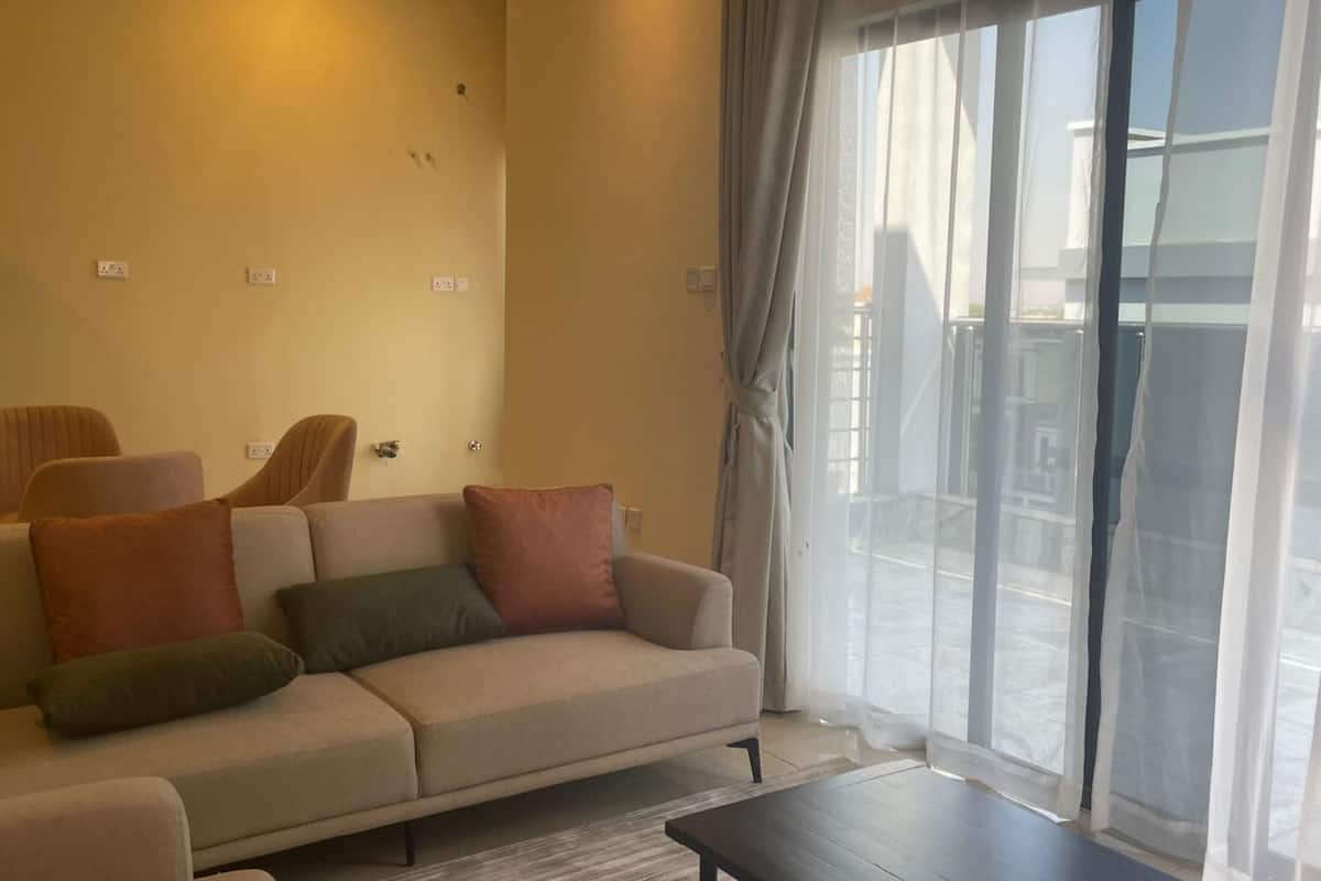 A living room with a beige sofa, green and brown cushions, a dark coffee table, and a large window with sheer curtains.