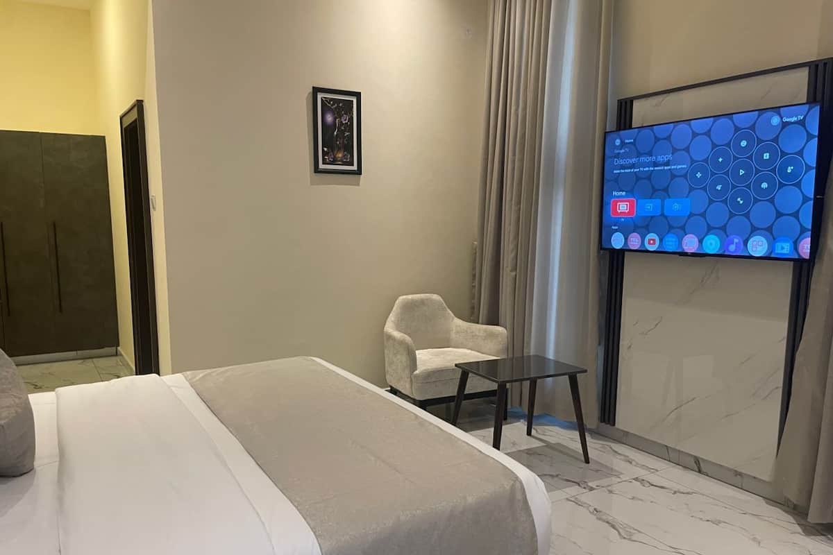 A modern hotel room with a large bed, a small armchair, a table, and a TV mounted on the wall displaying a home screen.
