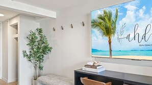 Et moderne værelse med et skrivebord, en bænk, en potteplante og et vægmonteret TV, der viser et strandbillede.