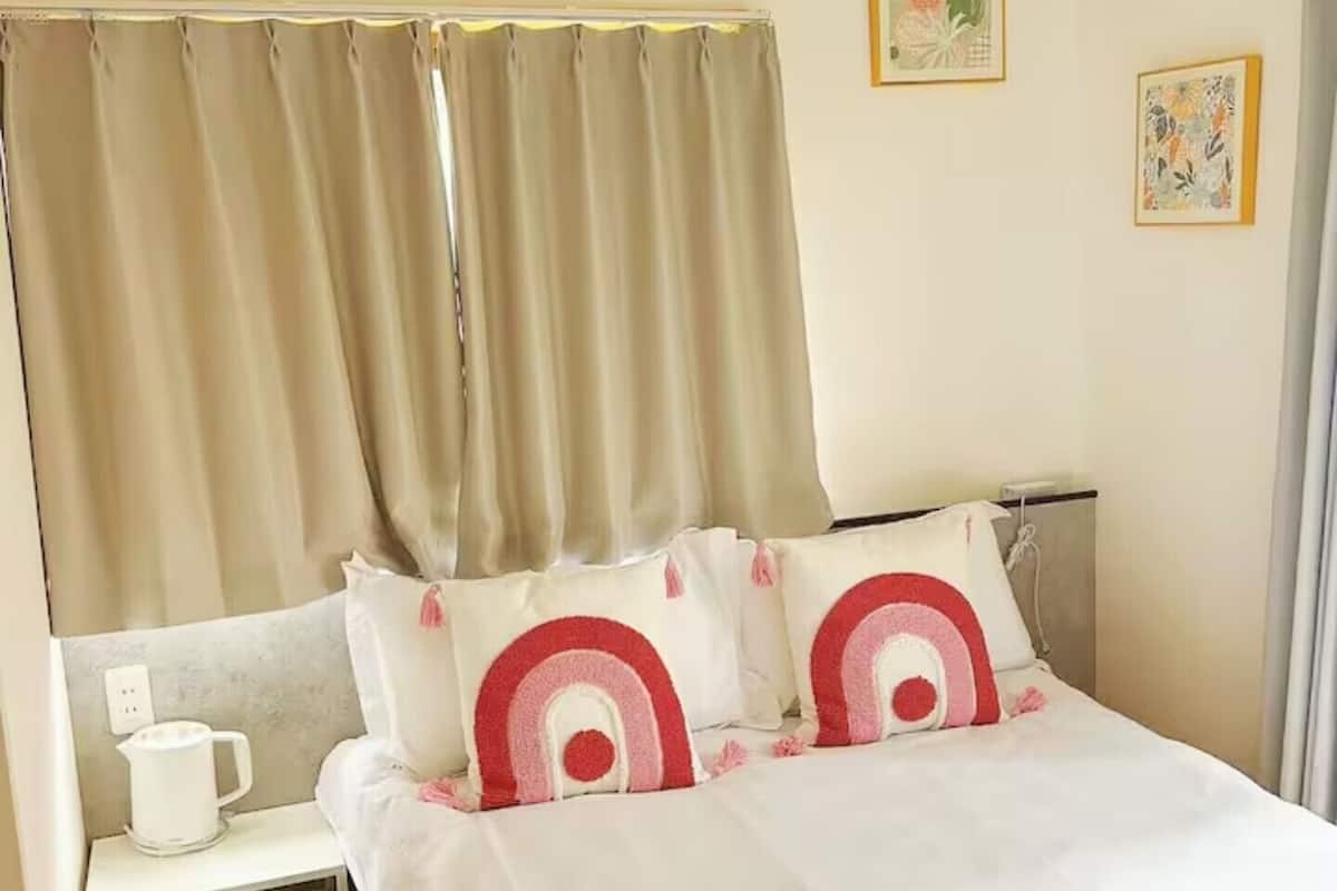 A neatly made bed with white bedding and two decorative pillows, a bedside table with a white mug, and a small drawer unit.