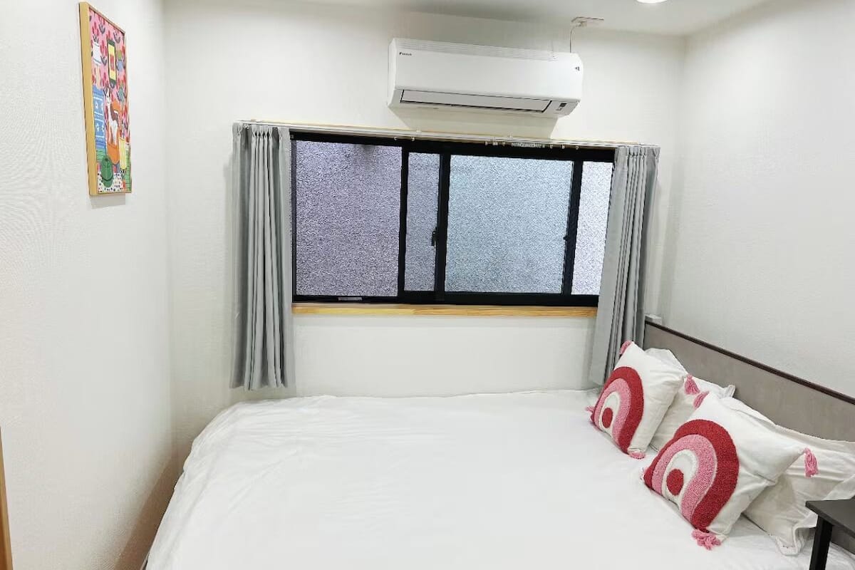 A single bed with white bedding, a window with grey curtains, an air conditioning unit above the window, and a small table beside the bed.