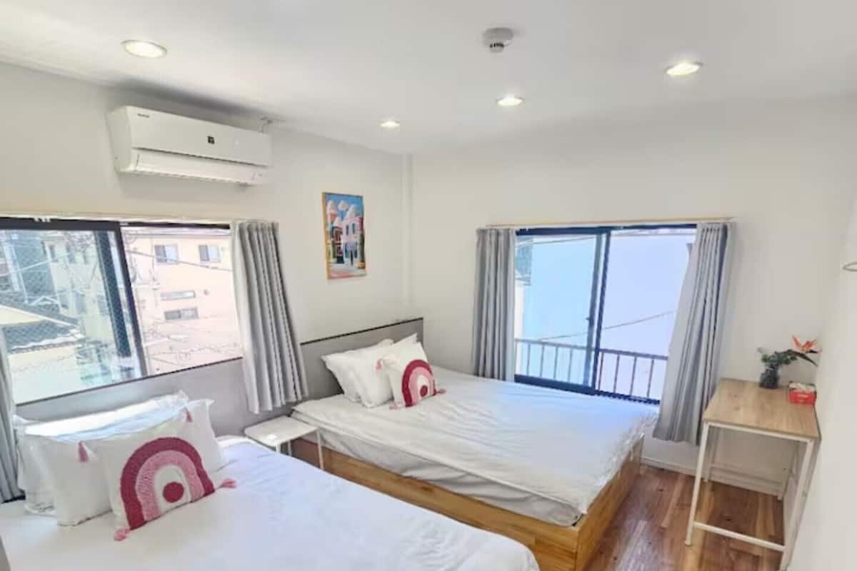 A hotel room with two beds, a small table, a window with curtains, and an air conditioning unit.