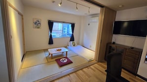 Une pièce avec un sol recouvert de tatamis, un futon servant de lit, une télévision et une petite table avec un coussin.