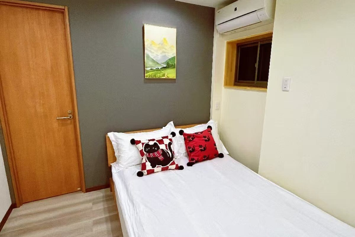 A bedroom with a bed, a door, a painting, and an air conditioning unit.
