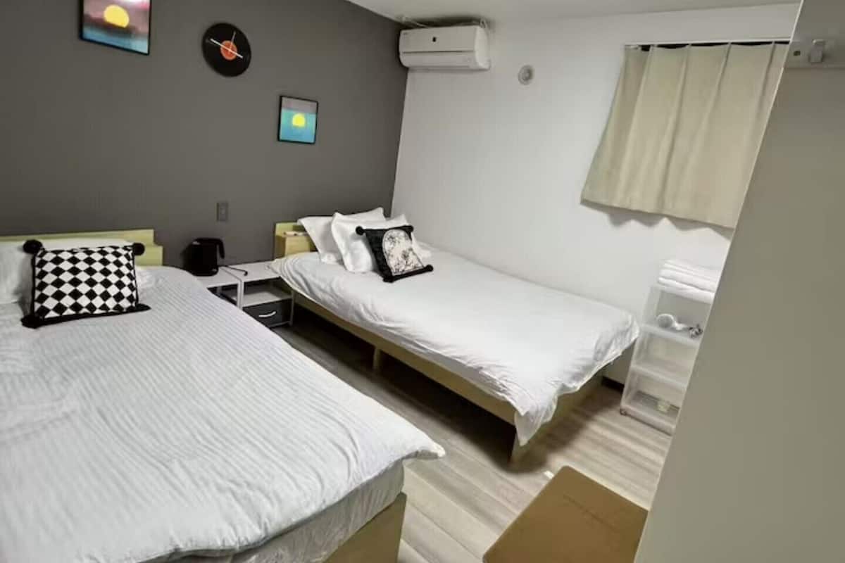 A room with two beds, a ceiling light, a wall clock, and a small window with curtains.