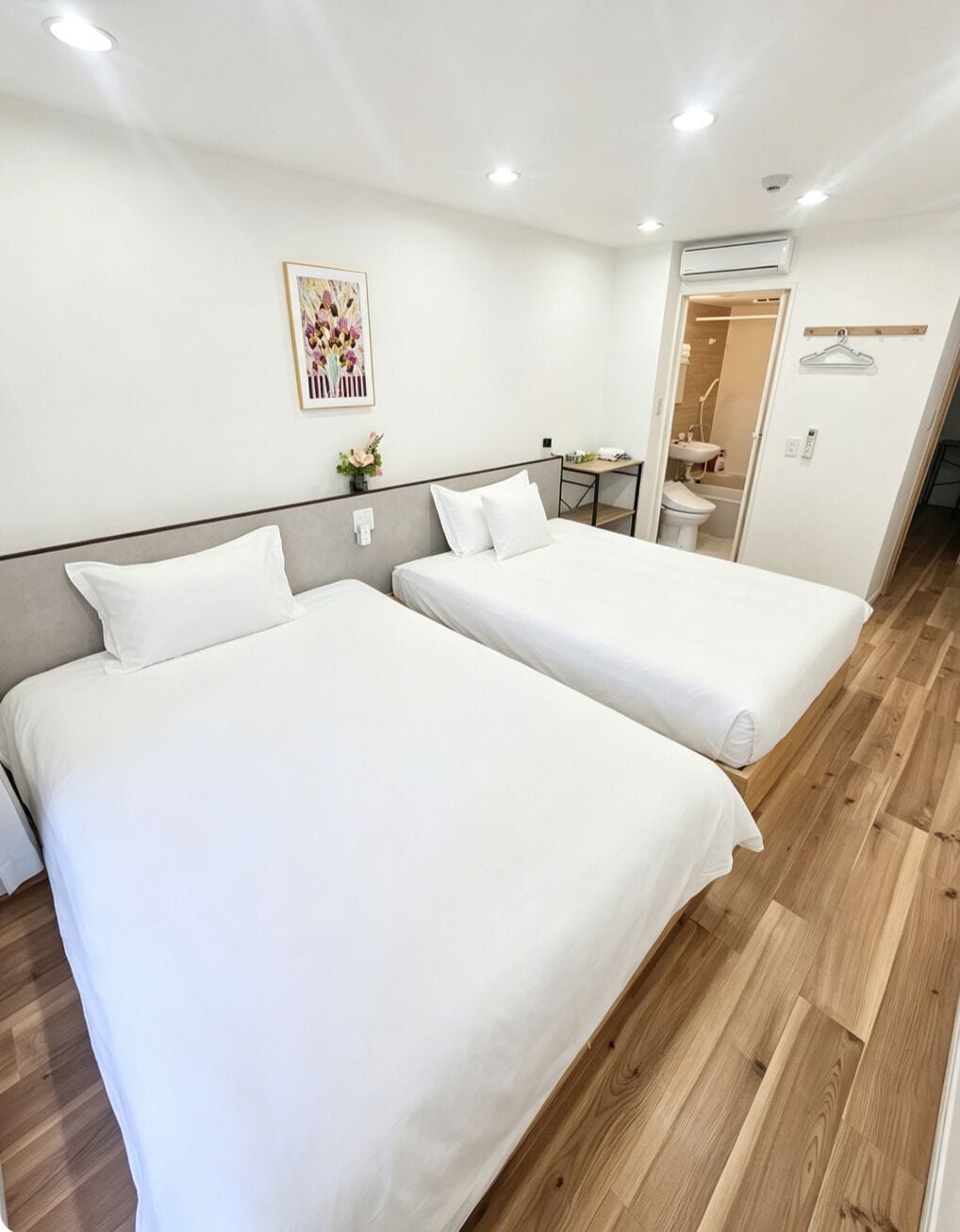 A hotel room with two beds, a small desk, a mirror, and a bathroom visible through an open door.