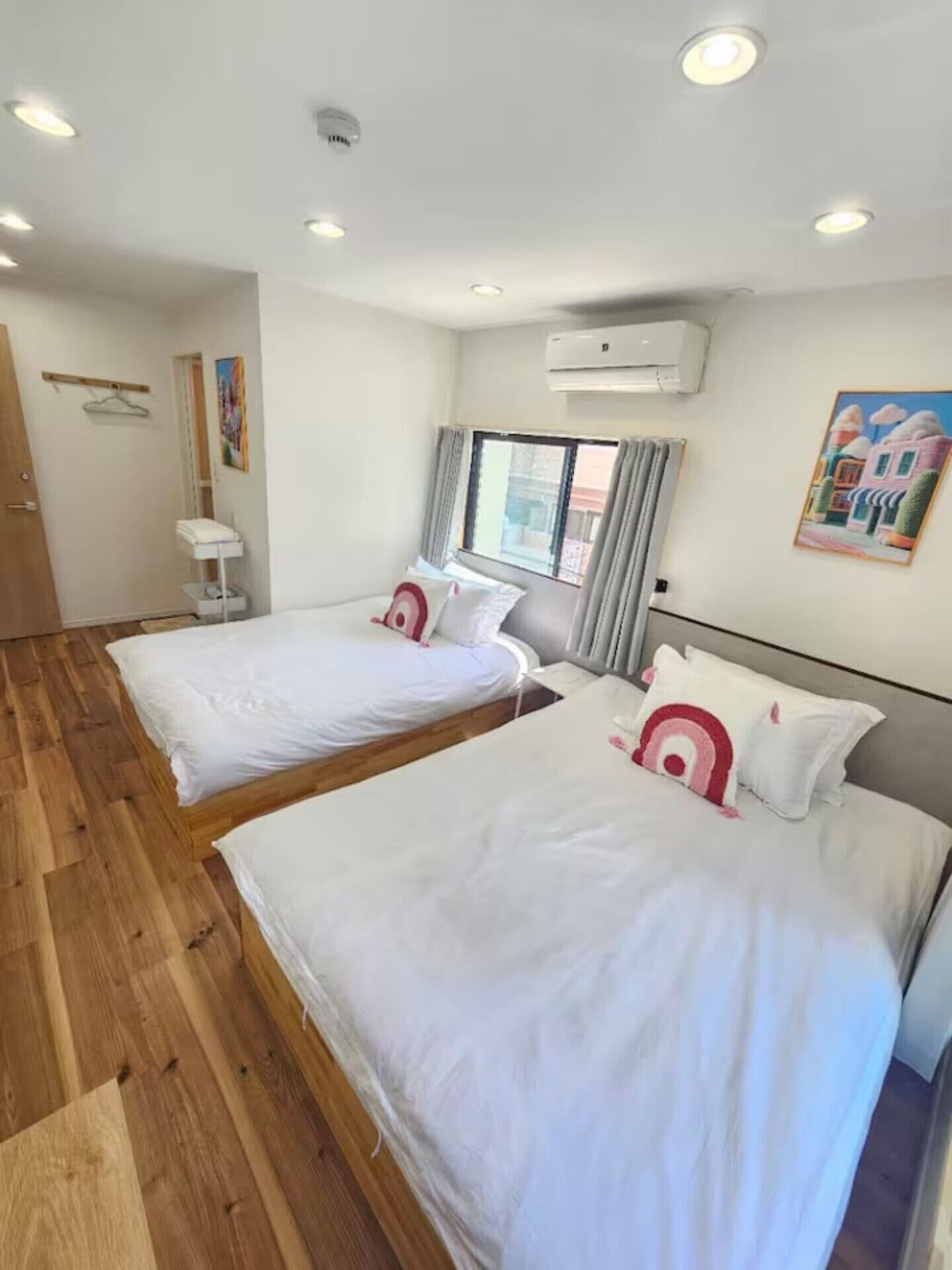 A hotel room with two beds, a wooden floor, an air conditioning unit, a window with curtains, and a painting on the wall.