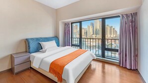 A hotel room with a large window, a bed with white bedding and an orange throw, a nightstand, and a view of a cityscape. - Shengmei Ya International Apartment  (Guangzhou)