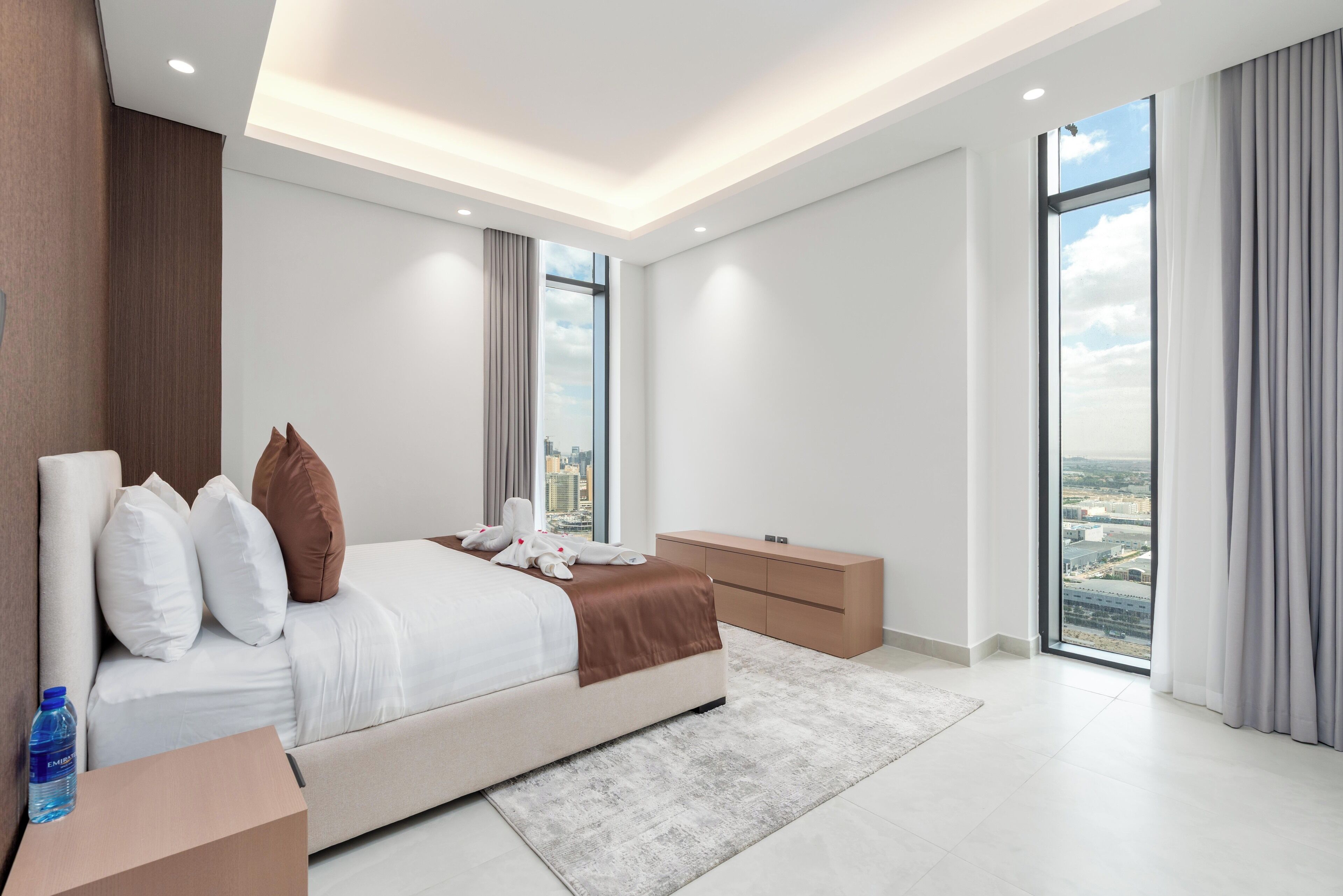A modern hotel room with a large bed, a wooden headboard, a city view, and a large window.