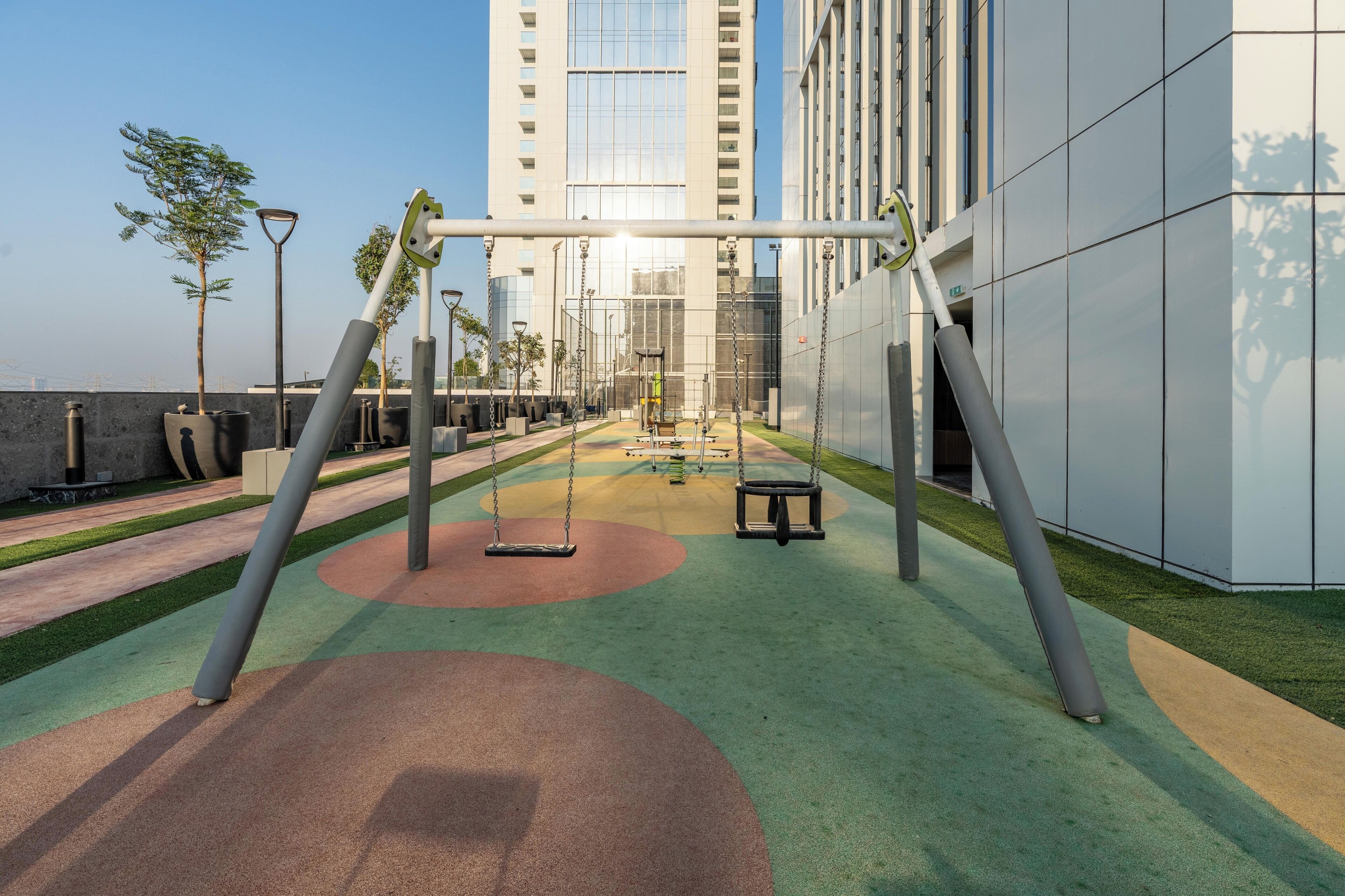A playground with swings and a slide, surrounded by a building with large windows and a paved area with green and brown surfaces.