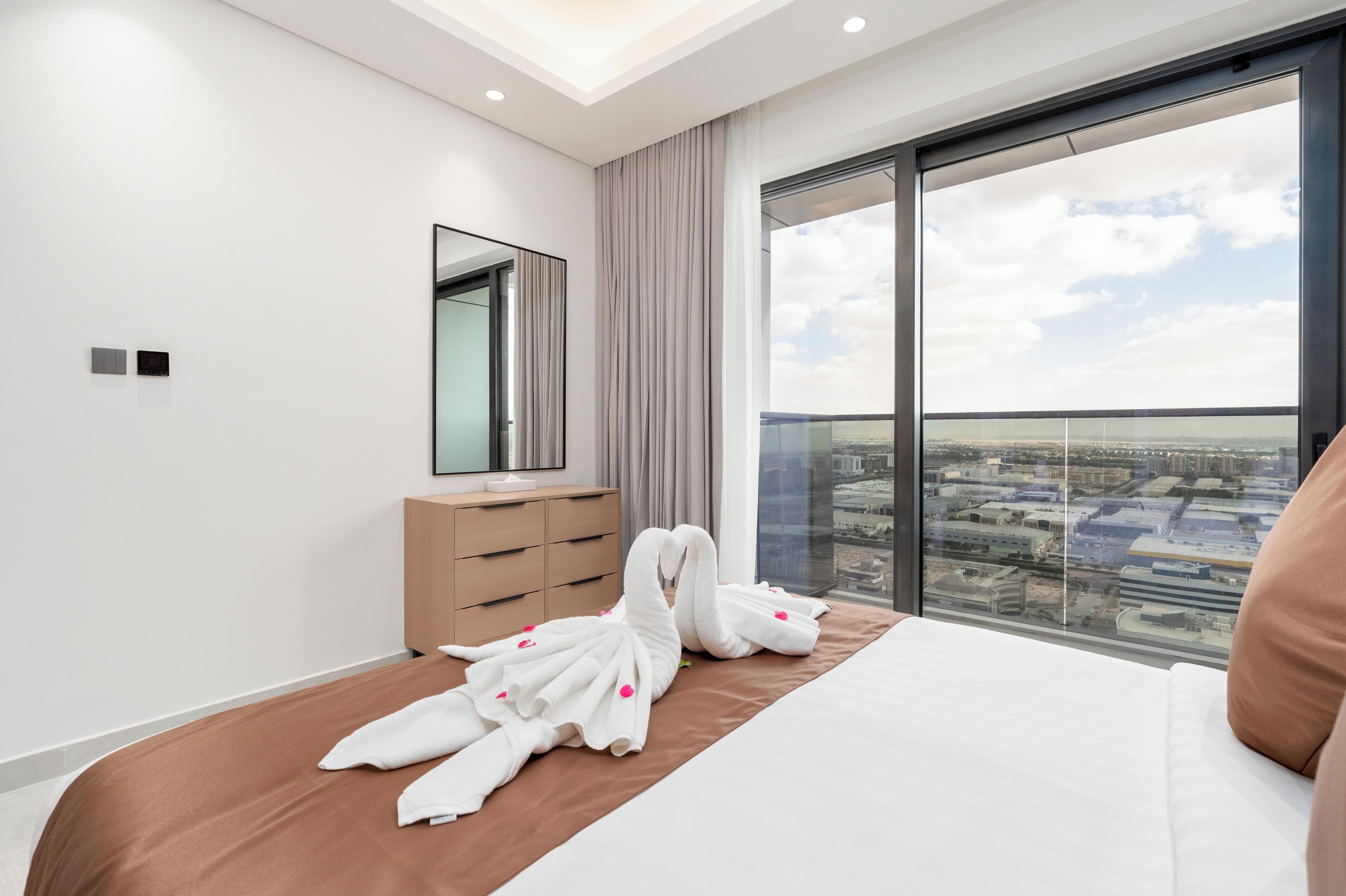 A modern hotel room with a large bed, a dresser, a mirror, and a city view through a large window.