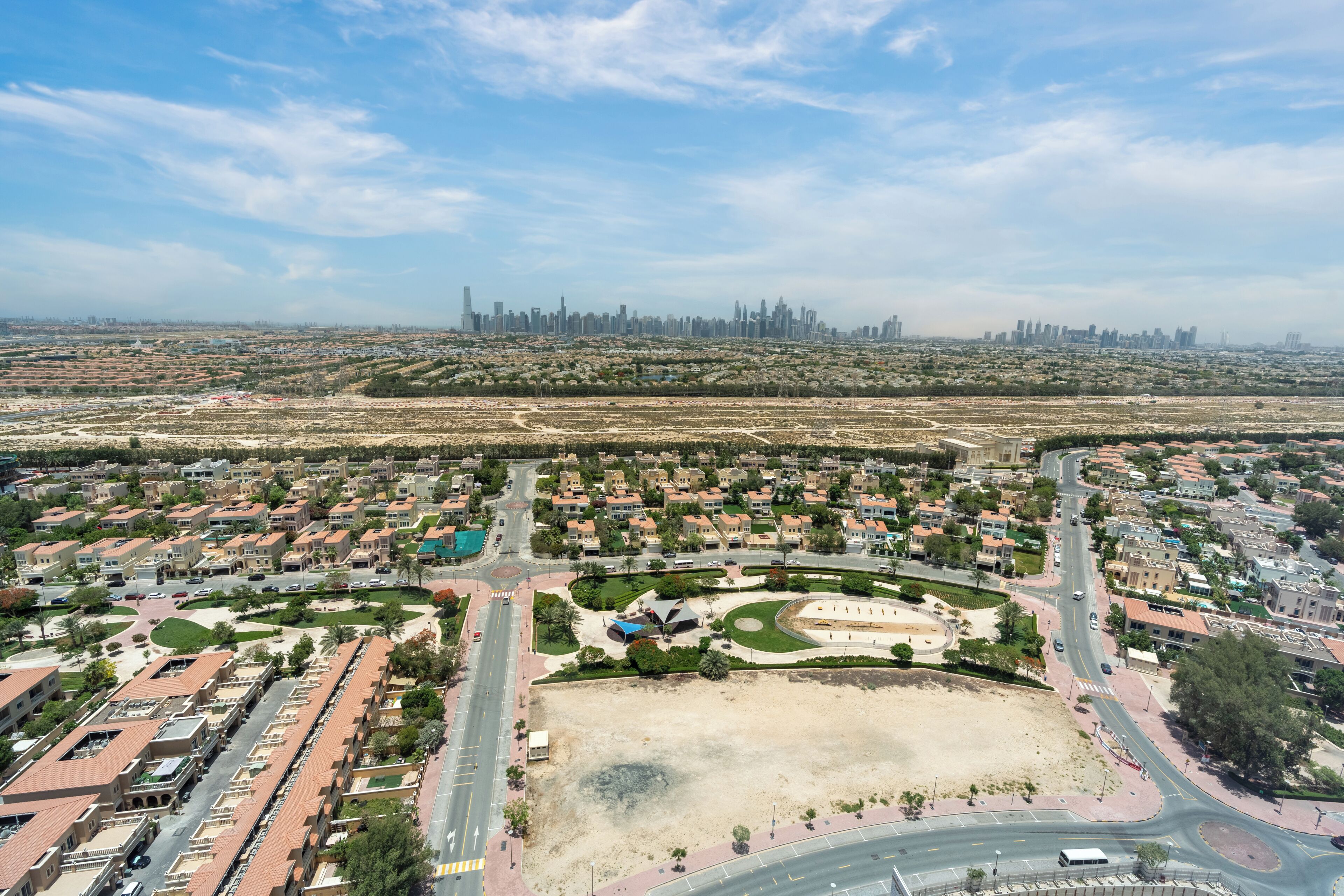 An aerial view of a residential area with a central park, a road network, and a distant city skyline.