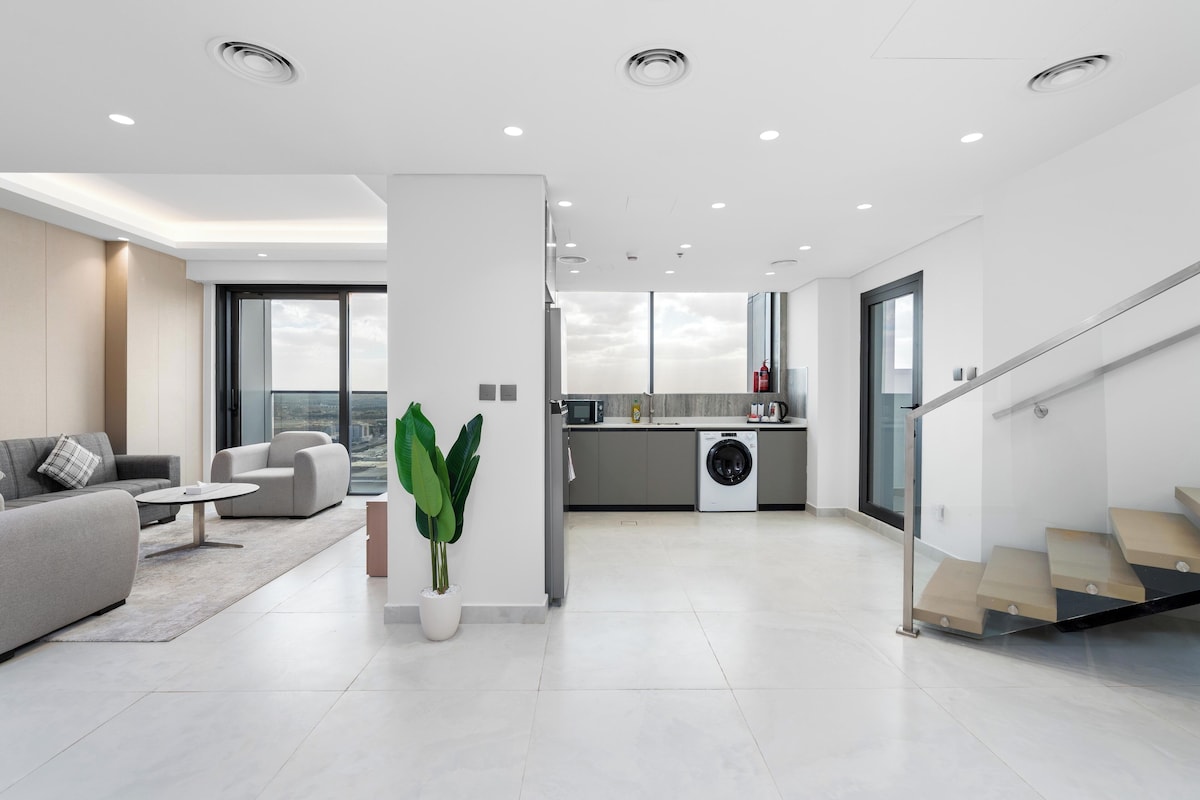 Una sala moderna con sofá, mesa de centro y planta en maceta. Se observa una zona de cocina con lavadora y una escalera.
