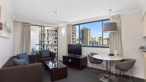 Una habitación de hotel moderna con sofá, mesa de centro, televisor, pequeña mesa de comedor y vista a la ciudad a través de amplias ventanas.