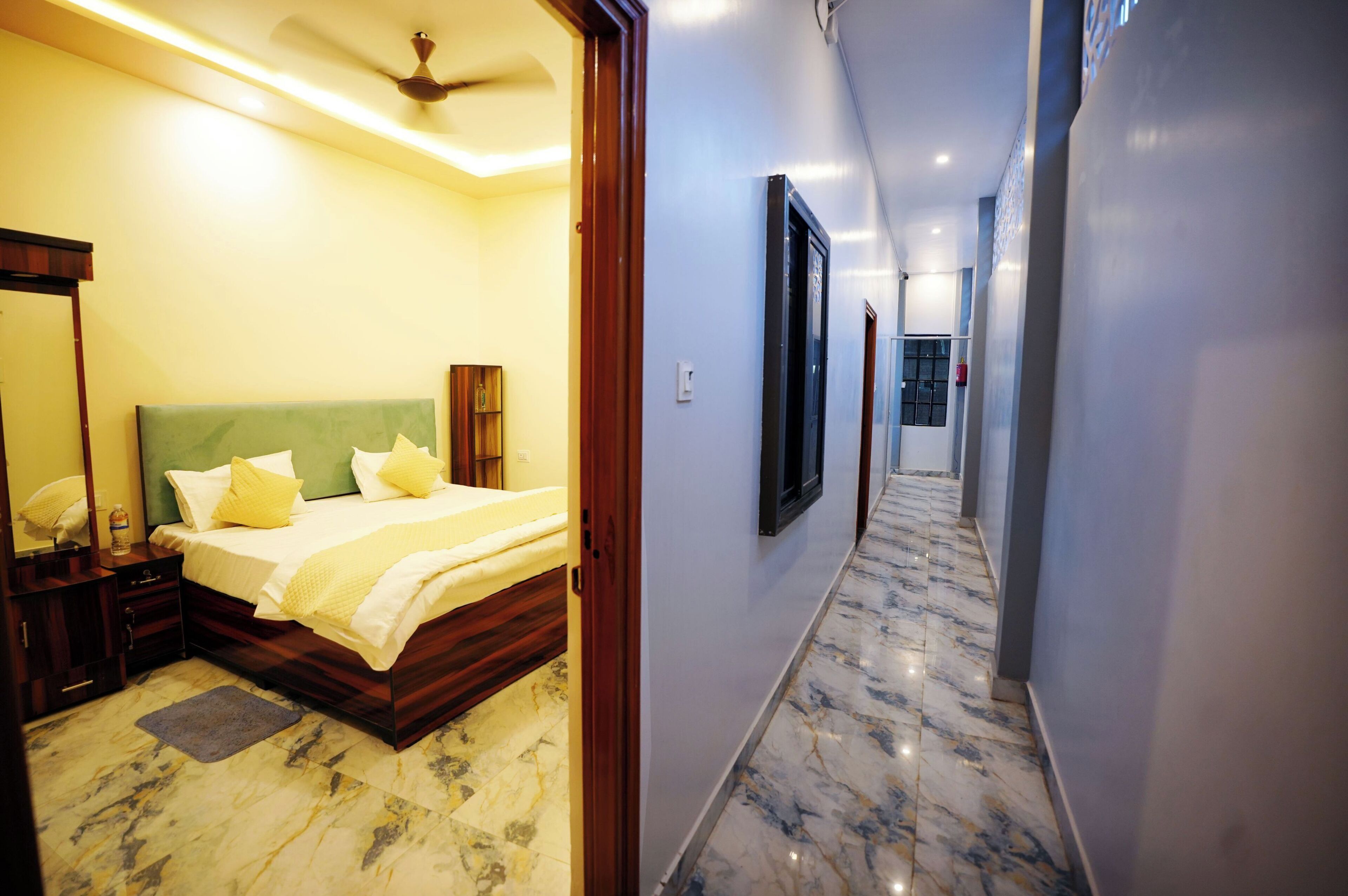 A hotel room with a bed, bedside table, and a mirror. A hallway with a marble floor leads to another room.